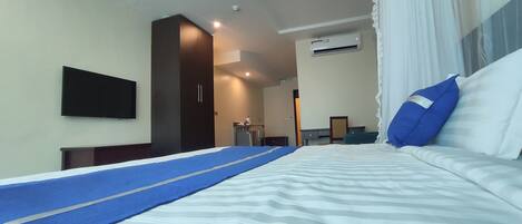 A hotel room with a bed, a TV mounted on the wall, a desk, and an air conditioning unit.