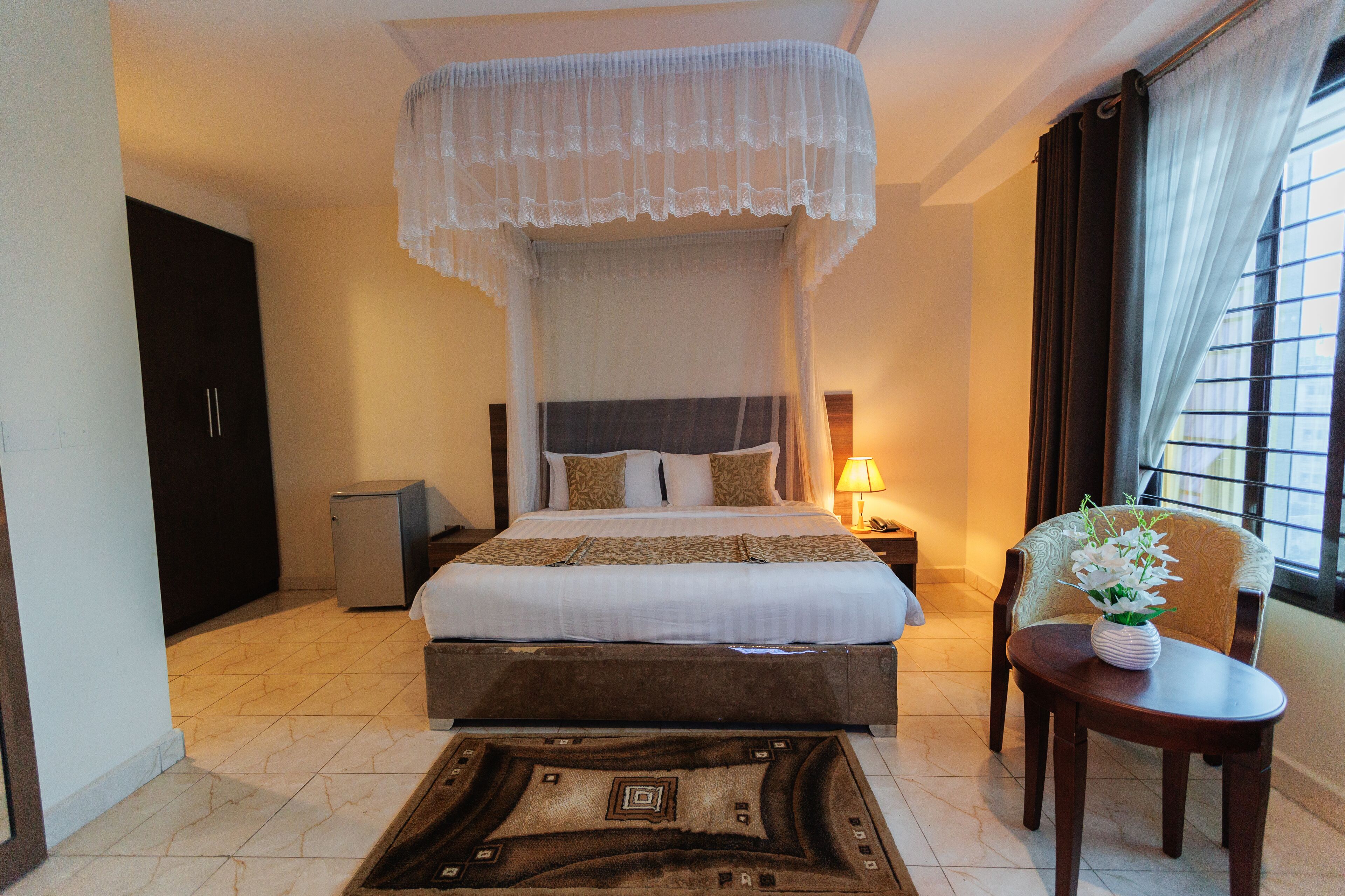 A hotel room with a bed, bedside table, lamp, chair, and a view of the city through the window.