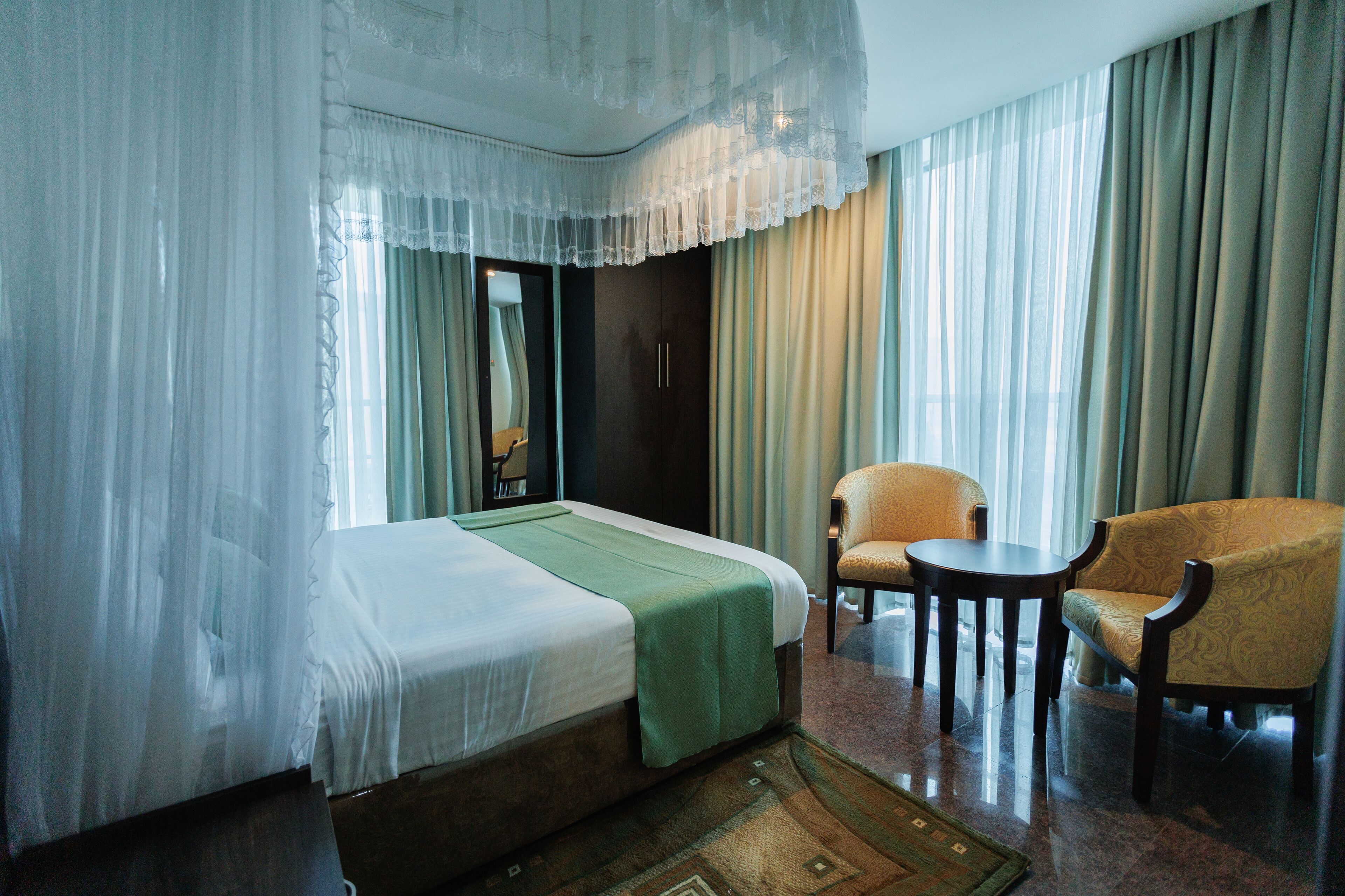 Habitación de hotel con cama, una manta verde, una silla, una mesita y cortinas traslúcidas.