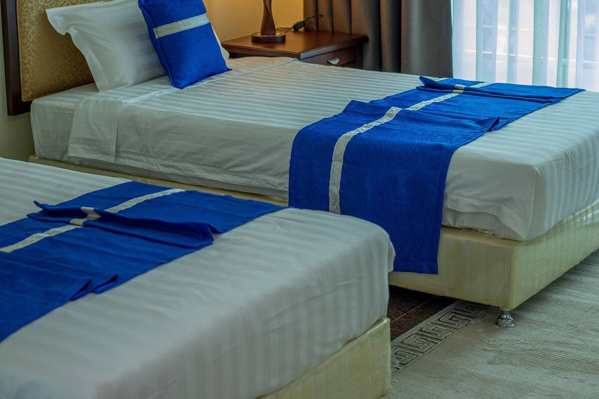 Habitación de hotel con dos camas, una mesita de noche con lámpara y vistas a la ciudad a través de amplios ventanales.