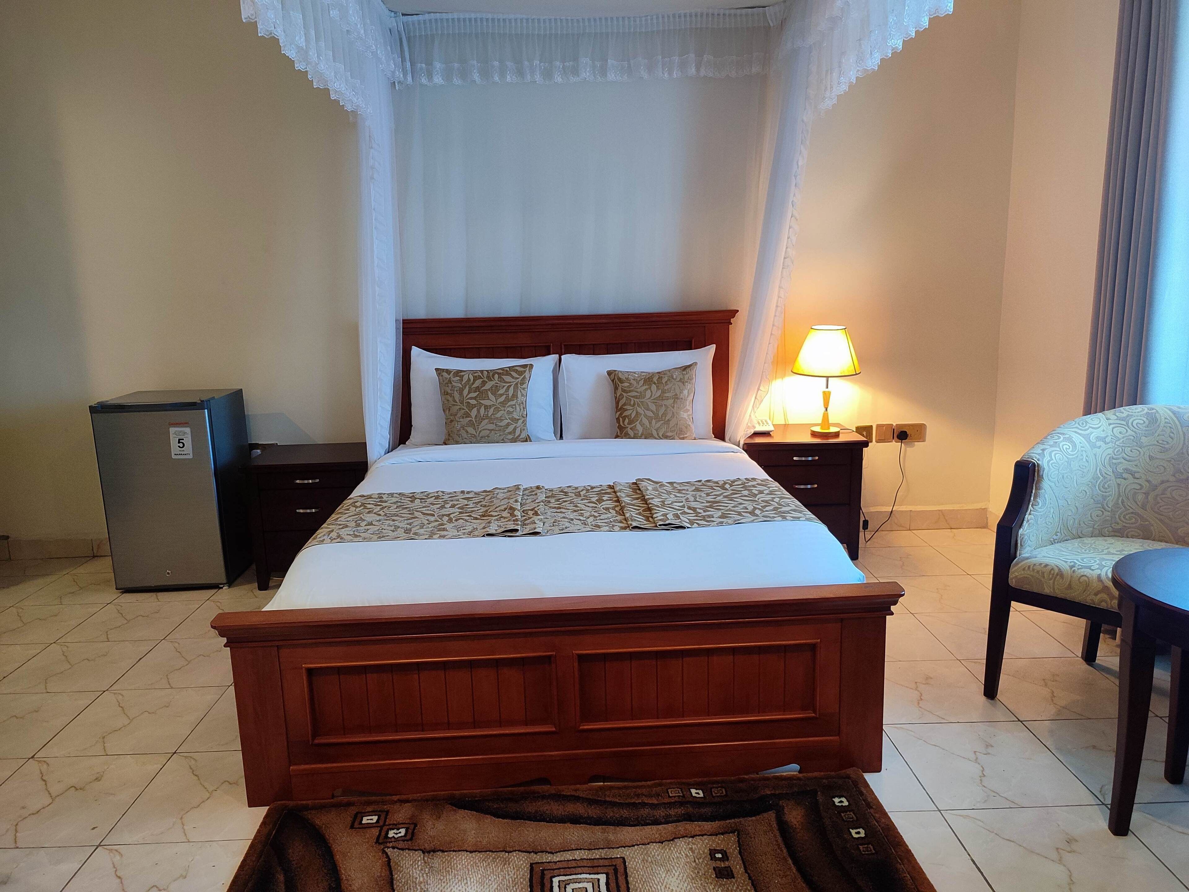 A neatly made bed with a wooden headboard, a bedside lamp, a chair, and a small refrigerator.