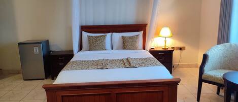 A neatly made bed with a wooden headboard, a bedside lamp, a chair, and a small refrigerator.