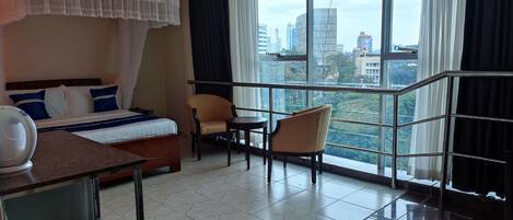 A hotel room with a bed, a chair, a table, and a large window offering a city view.