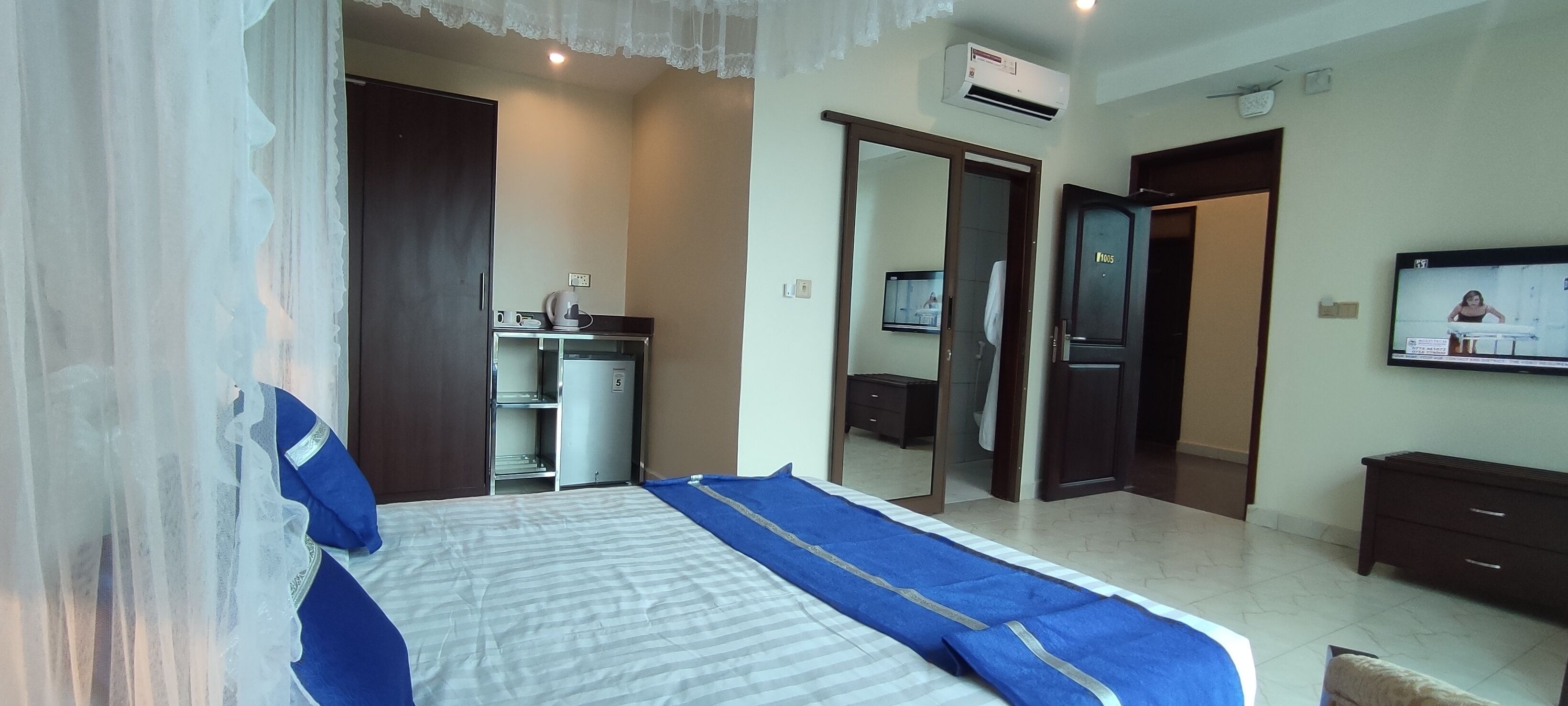 A hotel room with a bed, a wardrobe, a desk with a chair, a television, and an air conditioning unit.