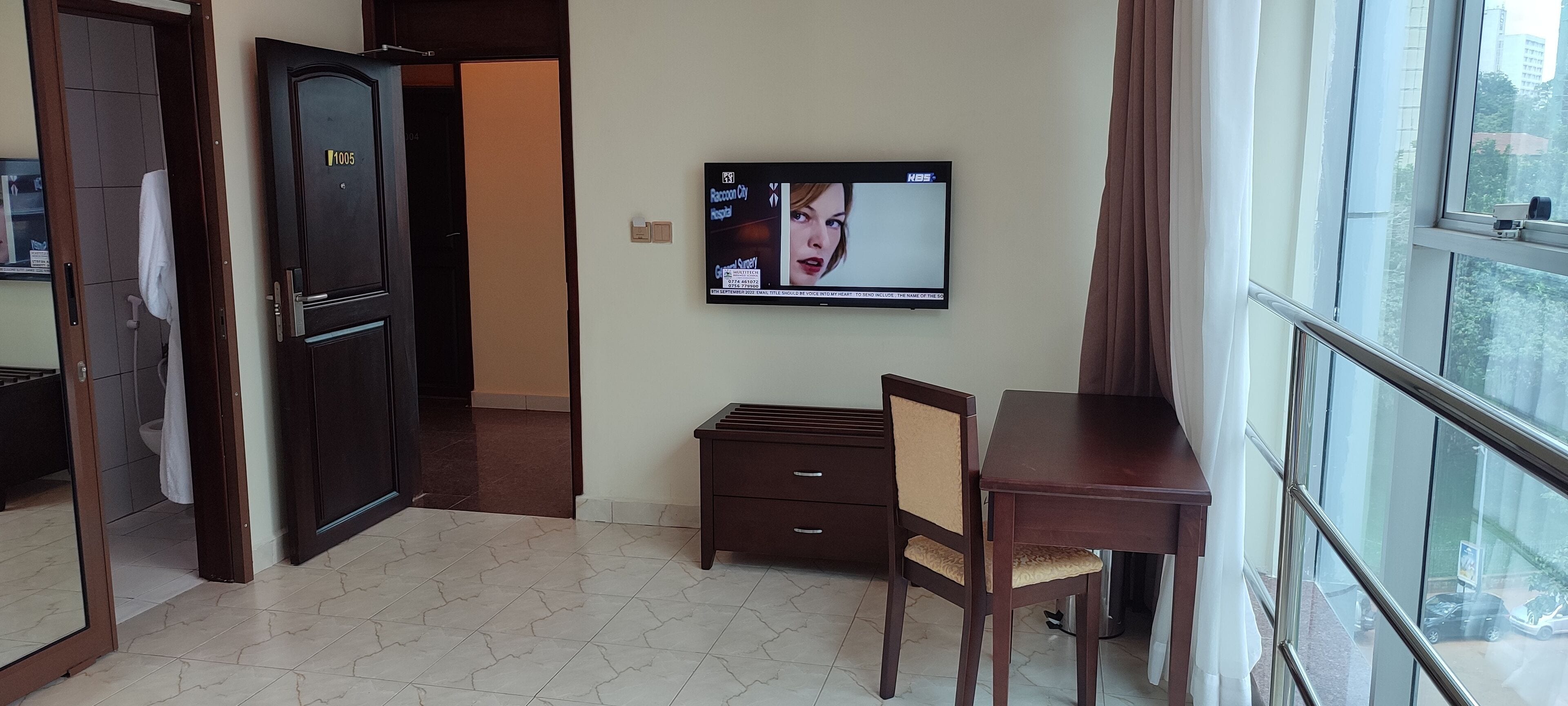 A hotel room with a wooden desk, a television mounted on the wall, and a large window with curtains.