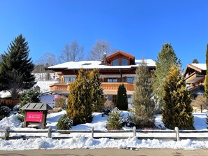 A multi-story building with a snow-covered roof, surrounded by evergreen trees and a wooden fence. - Country house Obermaiselstein THE VIEW - wonderful mountain view from apartment Besler (Obermaiselstein)