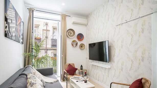 Una sala moderna con un sofá gris, una mesa de centro blanca, una silla de mimbre y un televisor de pantalla plana montado en la pared.