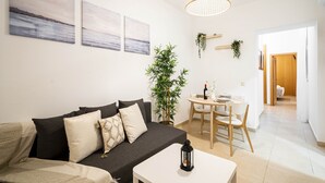 A modern living room with a dark grey sofa, a small white table, and a hanging plant. There are three framed artworks on the wall and a wooden door in the background.
