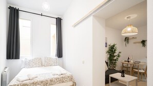 A bedroom with a bed, a bedside table, a small table with chairs, a hanging light fixture, and a window with curtains.