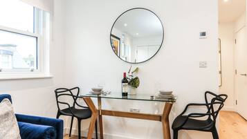 A modern dining area with a glass table, two black chairs, a round mirror, and a potted plant.