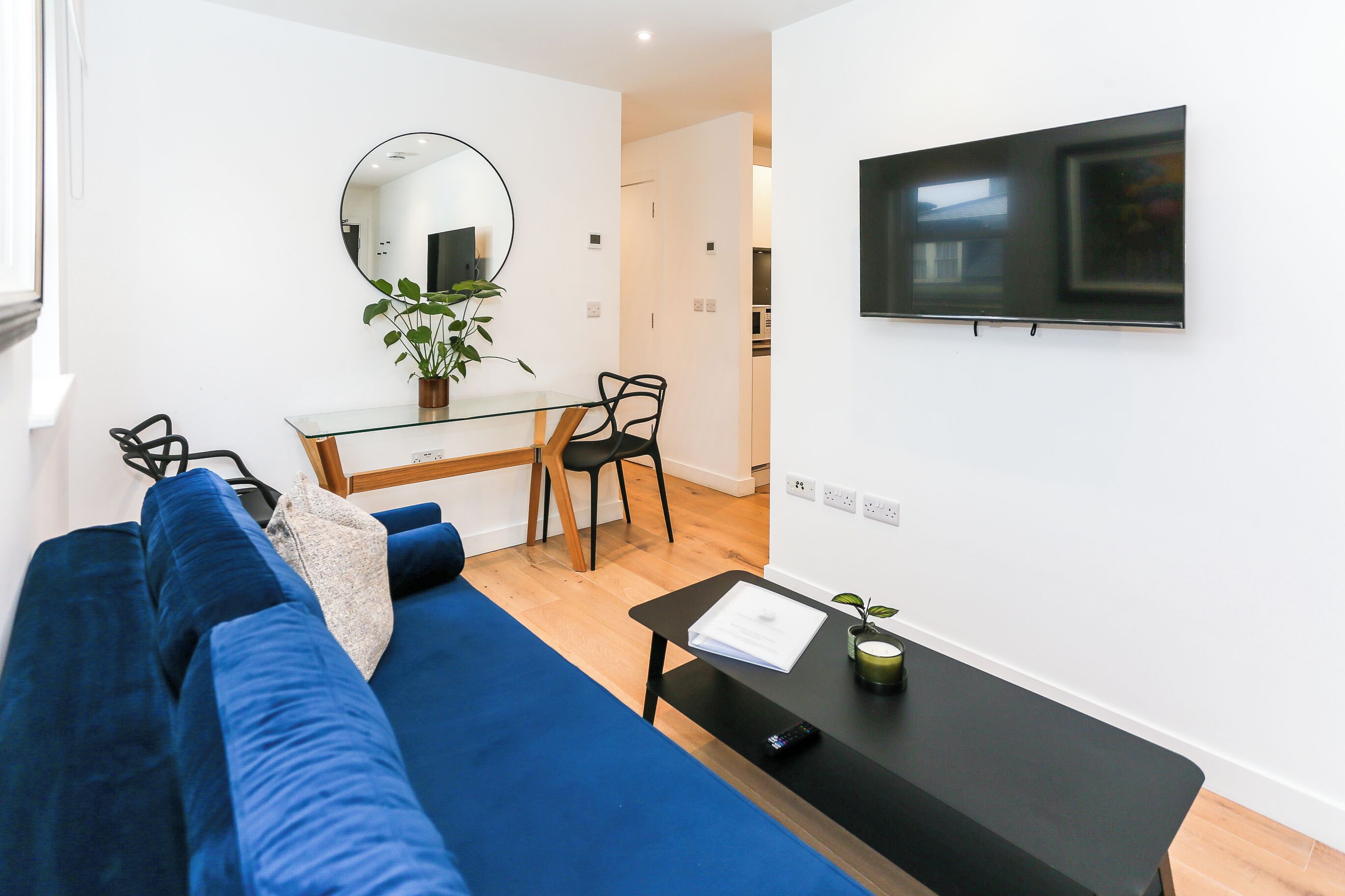 A modern living room with a blue sofa, a glass coffee table, a dining area with a chair, a small plant, and a TV mounted on the wall.