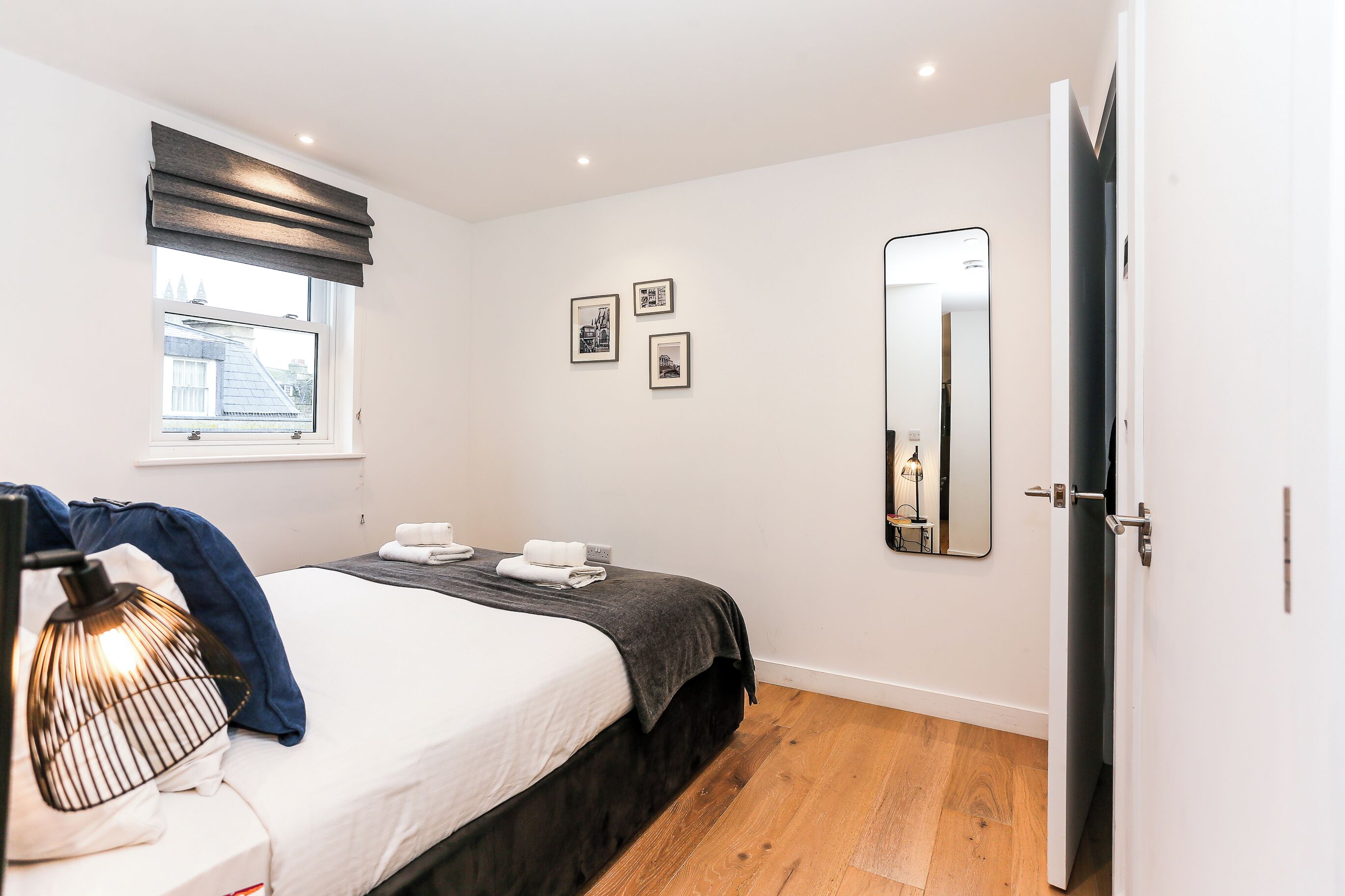 A modern hotel room with a neatly made bed, a bedside lamp, a window with blinds, and a mirror on the wall.