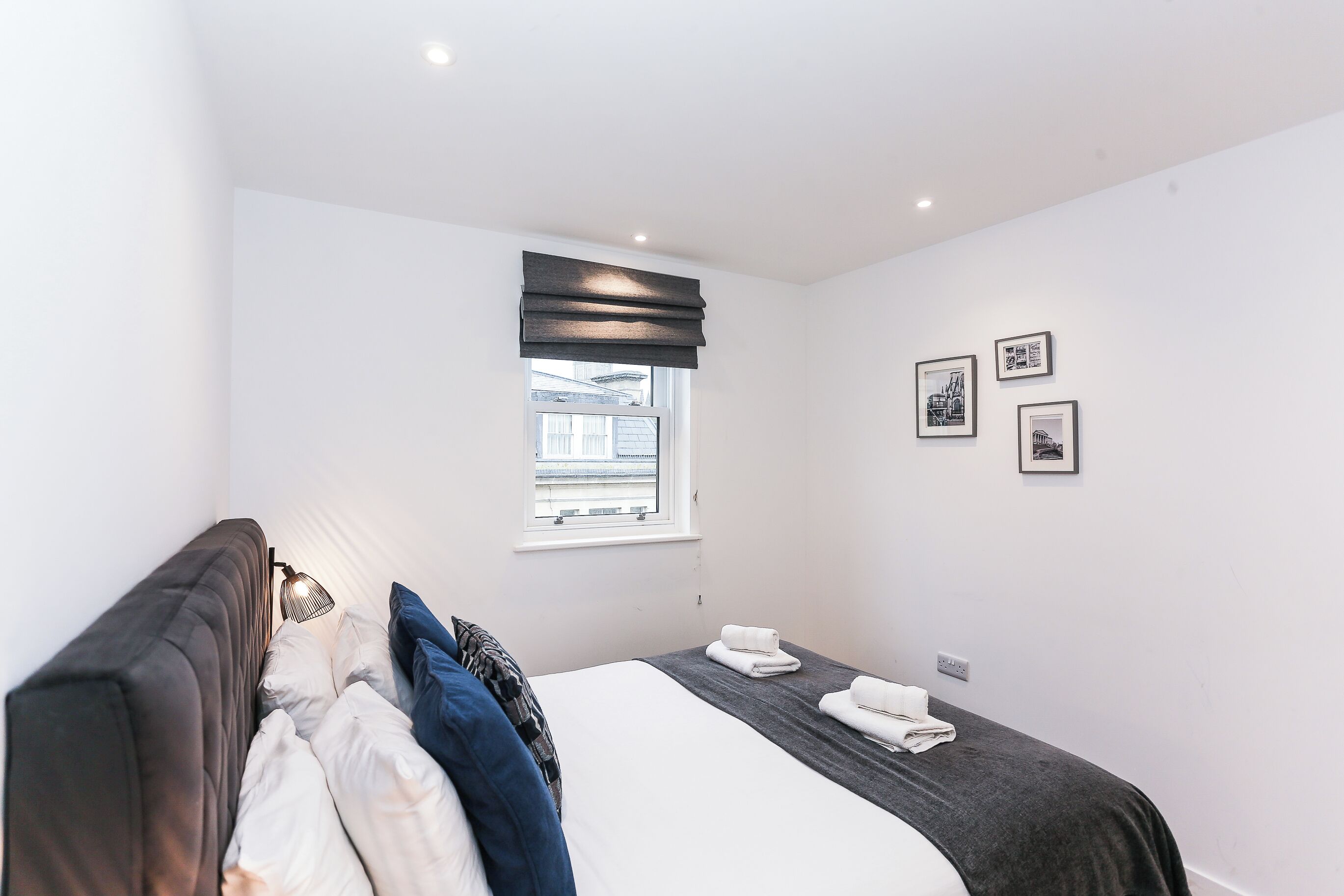 A modern bedroom with a large bed, a window with blinds, and a wall-mounted lamp.