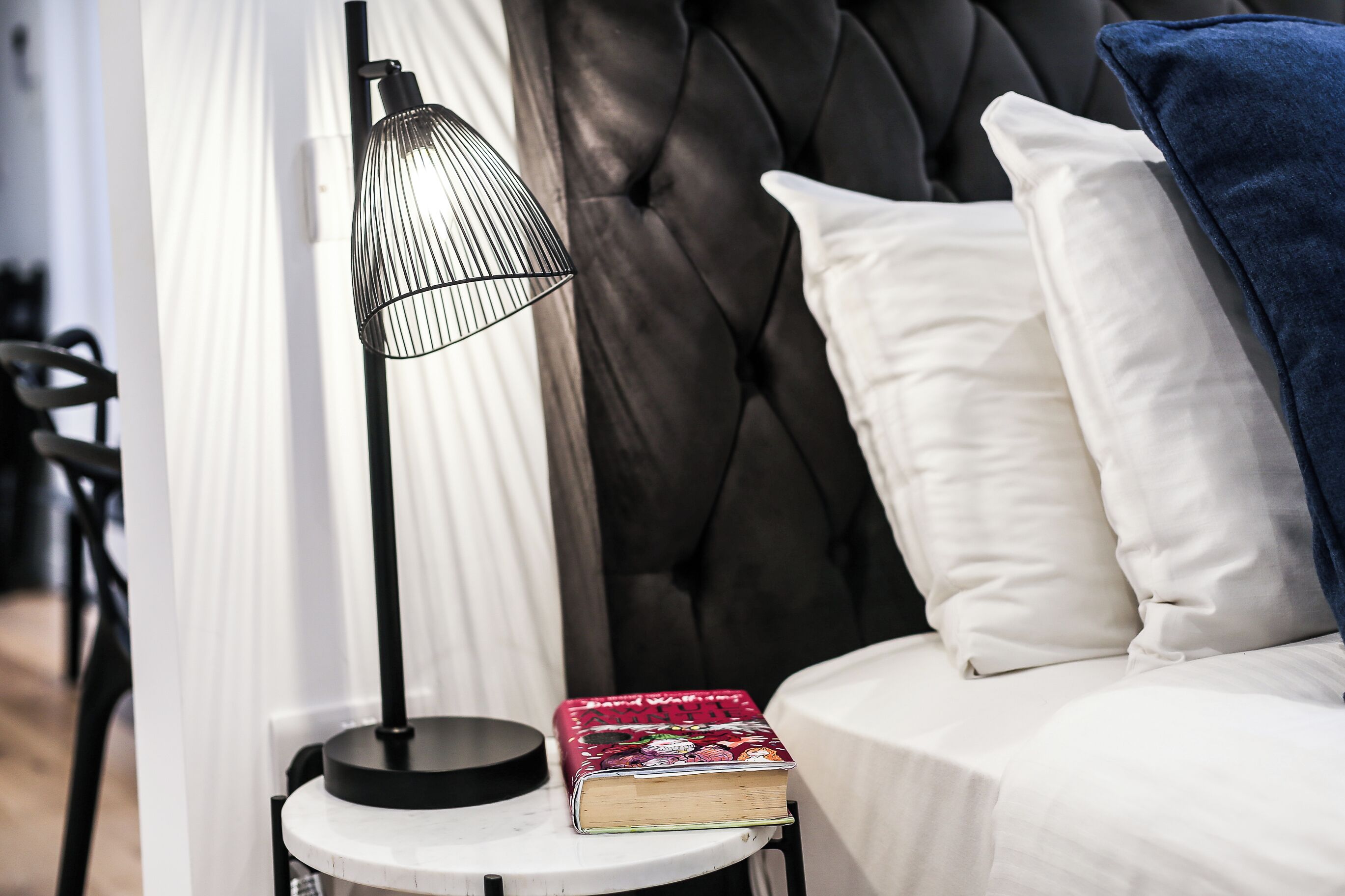 Ein Bett mit weißen und blauen Kissen, eine schwarze Lampe und ein Buch auf einem kleinen Tisch daneben.