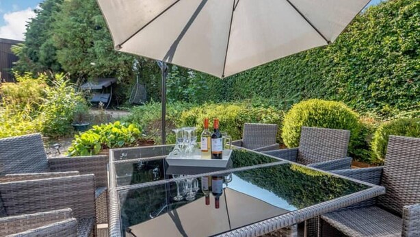 An outdoor patio with wicker furniture and a glass-top table, surrounded by well-maintained hedges and greenery. - Luxury 5-bed House With Tennis Court & Games Room (Steyning)