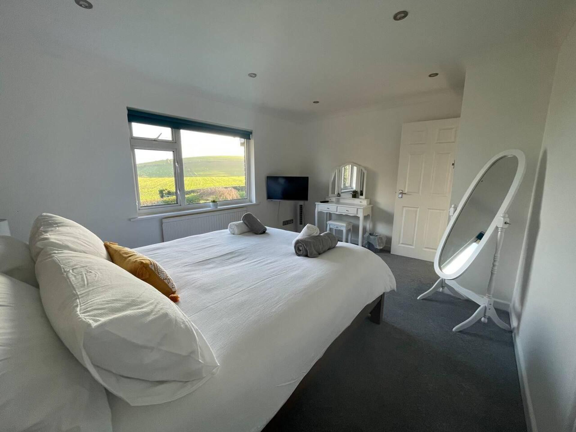 A bedroom with a large bed, a vanity with a mirror, a television, and a window with a view of fields.