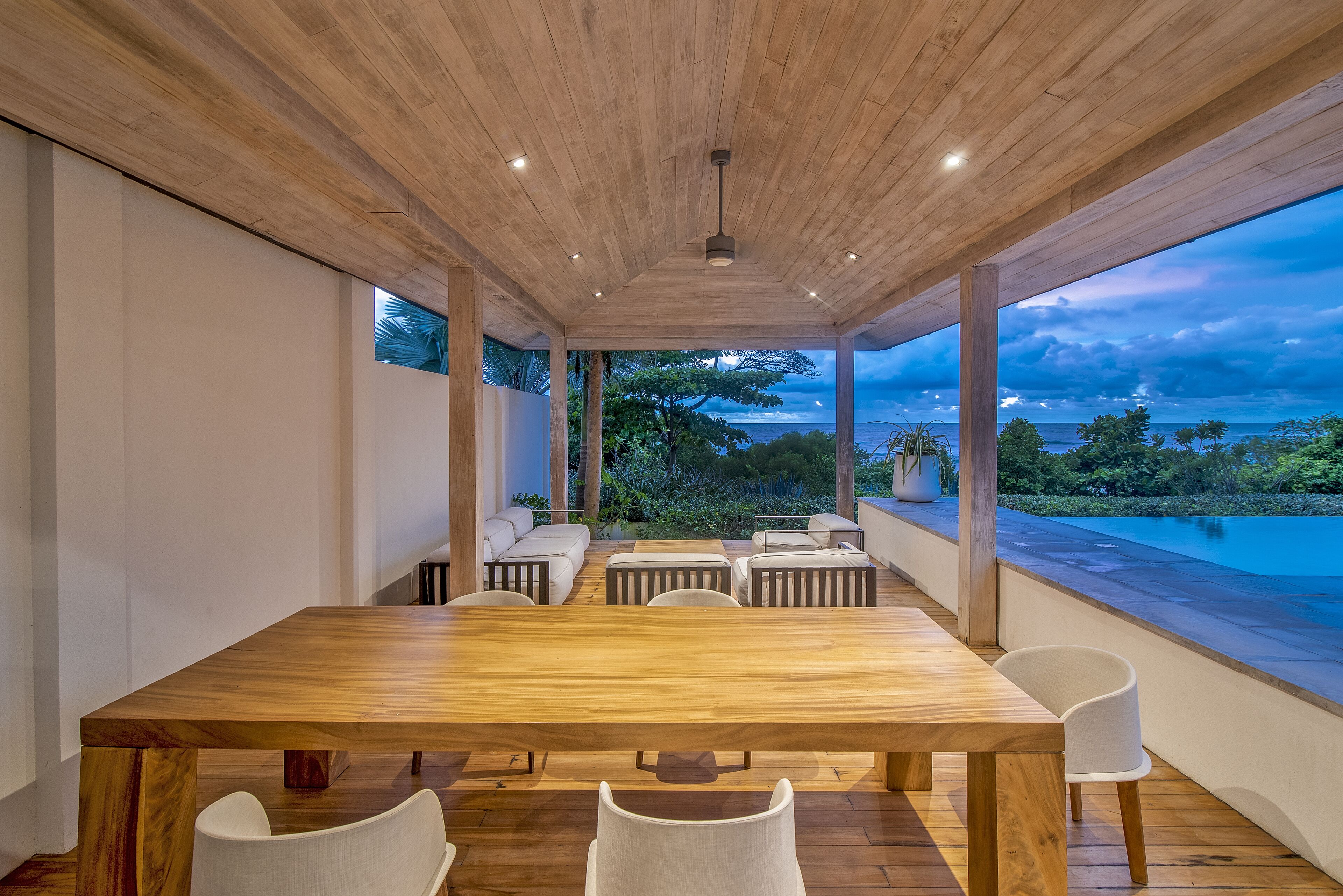 A spacious dining area with a wooden table and white chairs, overlooking a pool and lush greenery.