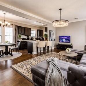 A modern living room with a grey sofa, a round coffee table, a TV, and a kitchen in the background.
