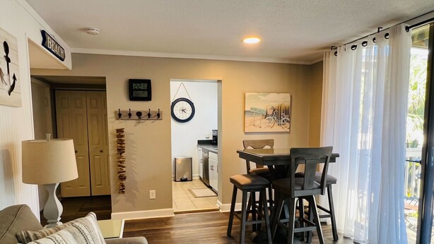 Una sala de estar con un sofá, una mesa de centro, una zona de comedor con una mesa y sillas, un reloj, un reloj de pared y una puerta que da a una cocina.
