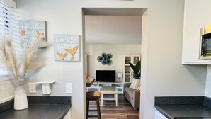 Una cocina moderna con piso de madera, gabinetes blancos y encimera negra. Cuenta con un comedor con mesa y sillas, un televisor y un adorno de pared.