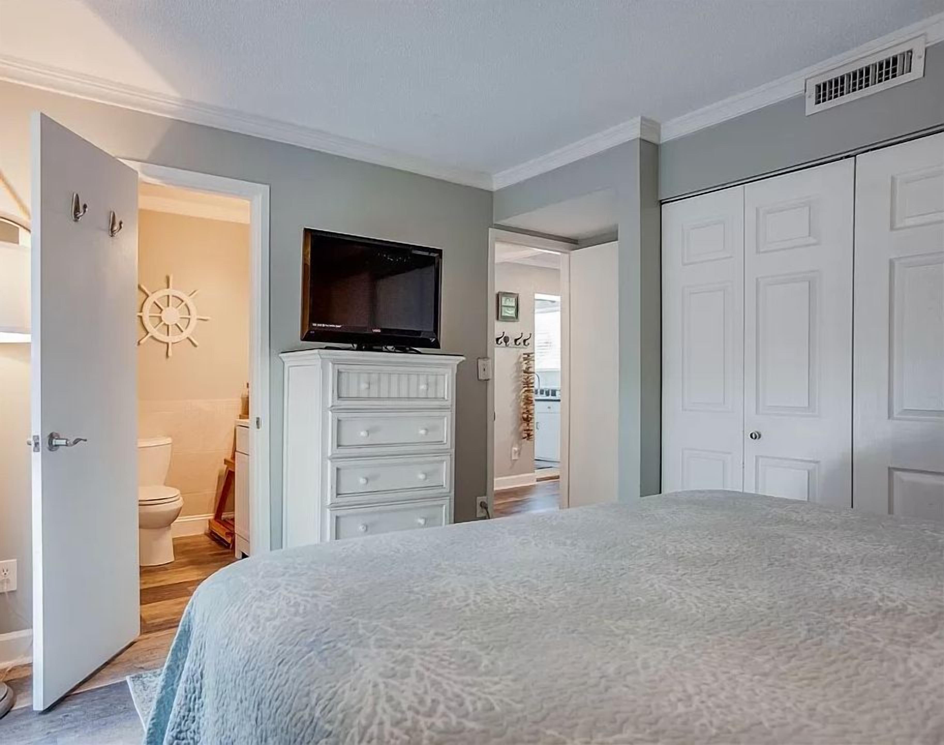 A bedroom with a bed, a television on a dresser, a wardrobe, and a bathroom visible through an open door.