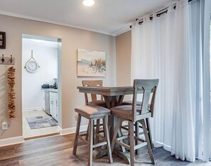 Een eethoek met een houten tafel en stoelen, een klok aan de muur, en een keuken op de achtergrond.