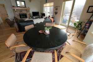 Un salon moderne avec une table ronde noire, des fauteuils en osier, une cheminée, un téléviseur et de grandes fenêtres.