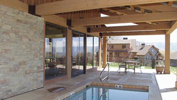 Piscina al aire libre con pared de piedra y vigas de madera, rodeada de puertas de cristal y un patio cubierto.