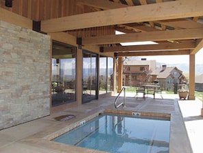 Une piscine extérieure avec un mur en pierre et des poutres en bois, entourée de portes vitrées et d’un patio couvert.