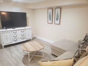 Uma sala de estar com uma TV de tela plana, um armário branco com gavetas, uma mesa de centro redonda de madeira, um sofá bege e duas gravuras botânicas emolduradas na parede.