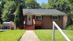 A single-story brick house with a front porch, a small shed to the left, and a metal railing in the foreground. - Amazing Escape - 4 BEDS, 3 BEDROOMS, Private Deck near DC (Annandale)