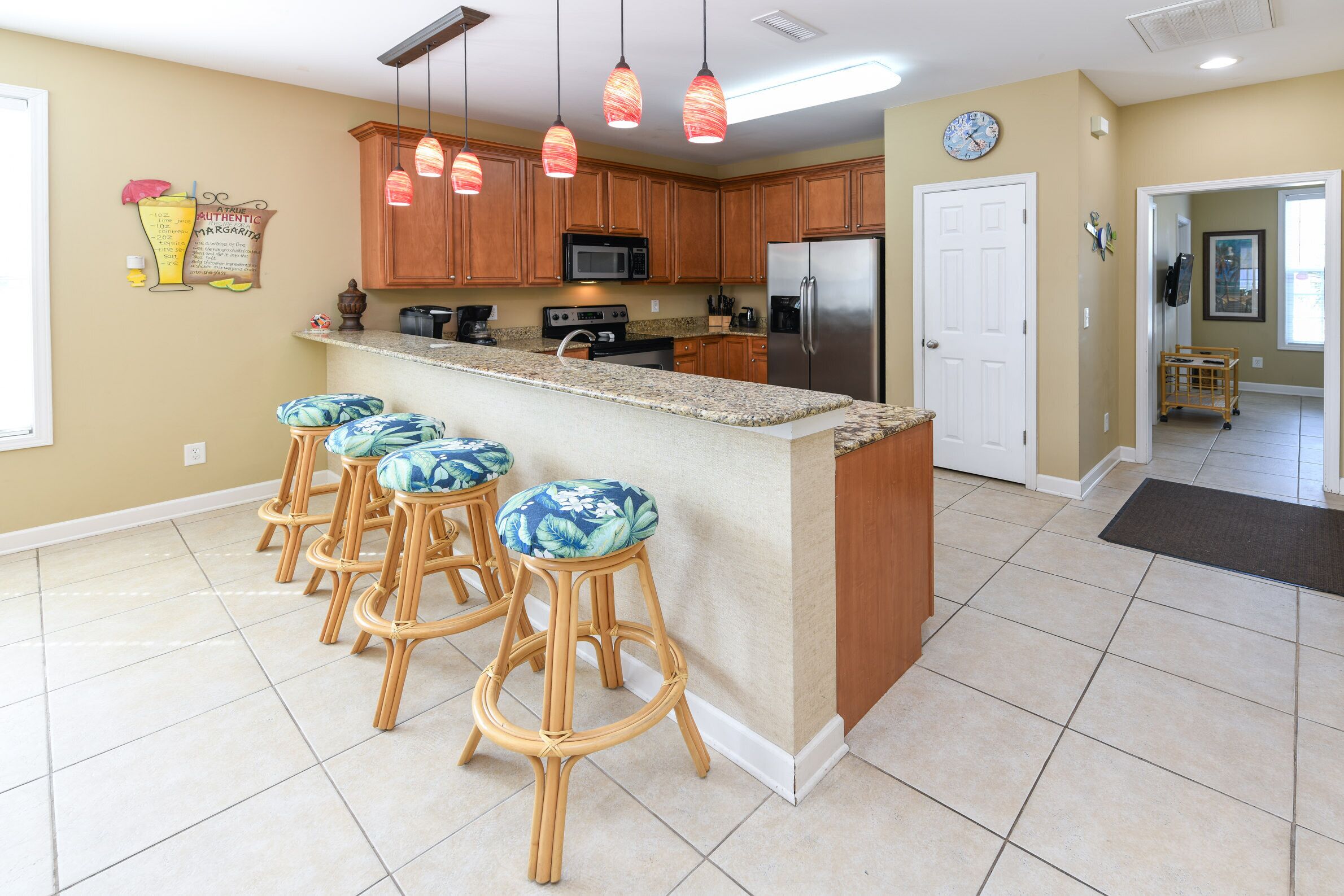 A kitchen with wooden cabinets, a stainless steel refrigerator, a granite countertop, and four bar stools with blue floral cushions.