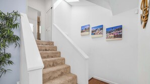 Un escalier dont les marches sont recouvertes d’une moquette beige, menant à un palier aux murs blancs. Trois tableaux encadrés sont accrochés au mur au-dessus de l’escalier.
