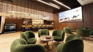 Un espace lounge moderne avec des fauteuils à revêtement vert, une table basse ronde et un bar doté d’un mur en bois et d’un grand écran diffusant une scène de désert.