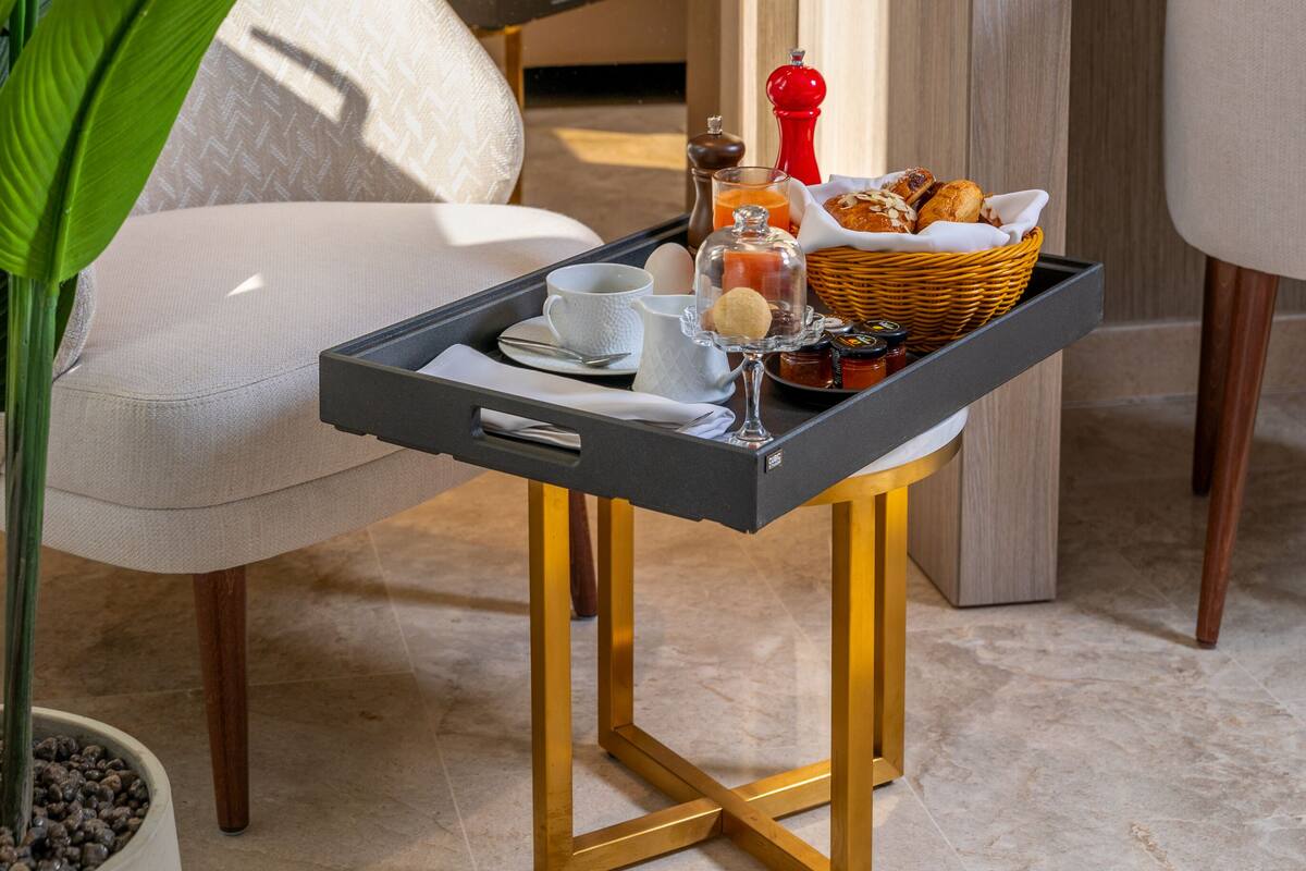 A hotel room with a bed, a bedside table with a vase of flowers, a tray with breakfast items, and a potted plant.