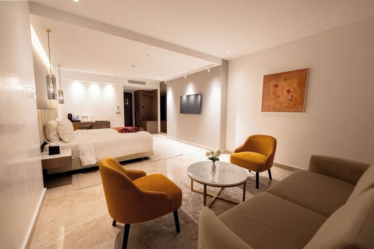 A modern hotel room with a bed, two armchairs, a coffee table, and a TV.