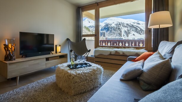Un salon moderne doté d’un téléviseur à écran plat, d’un pouf moelleux, d’un canapé avec des coussins et d’une grande fenêtre offrant une vue sur les montagnes.