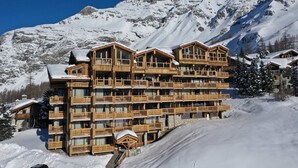 Un chalet en bois à plusieurs étages, doté de balcons, qui se dresse sur fond de montagnes enneigées.