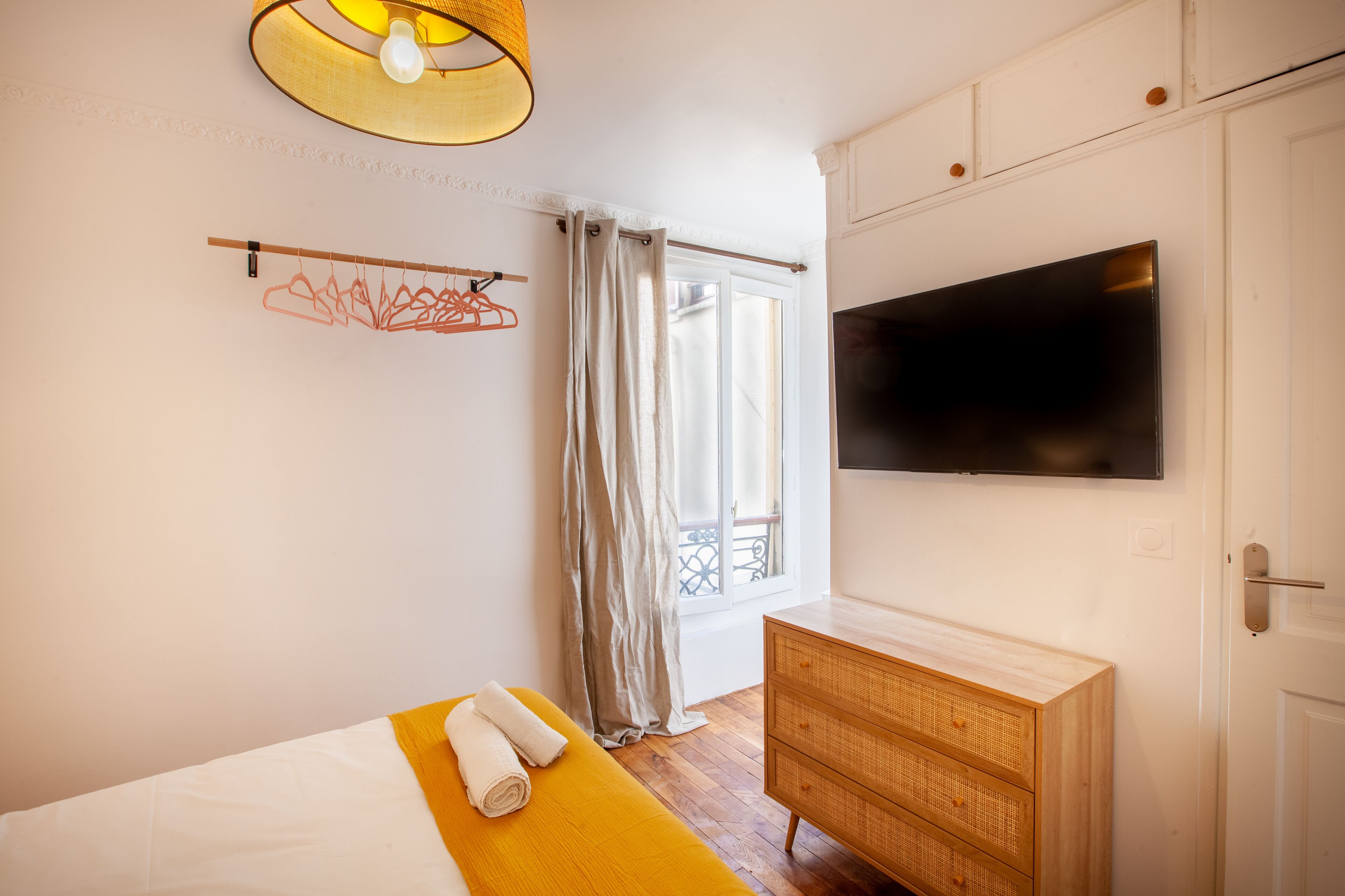 A bedroom with a bed, a wooden dresser, a television on the wall, and a window with curtains.