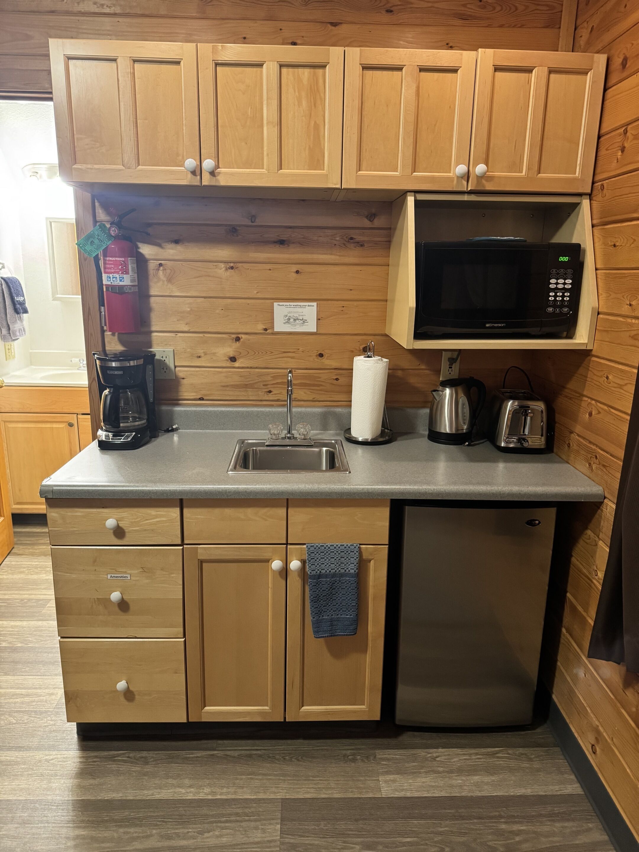 A compact kitchen with wooden cabinets, a stainless steel sink, a microwave, a coffee maker, a toaster, and a kettle.