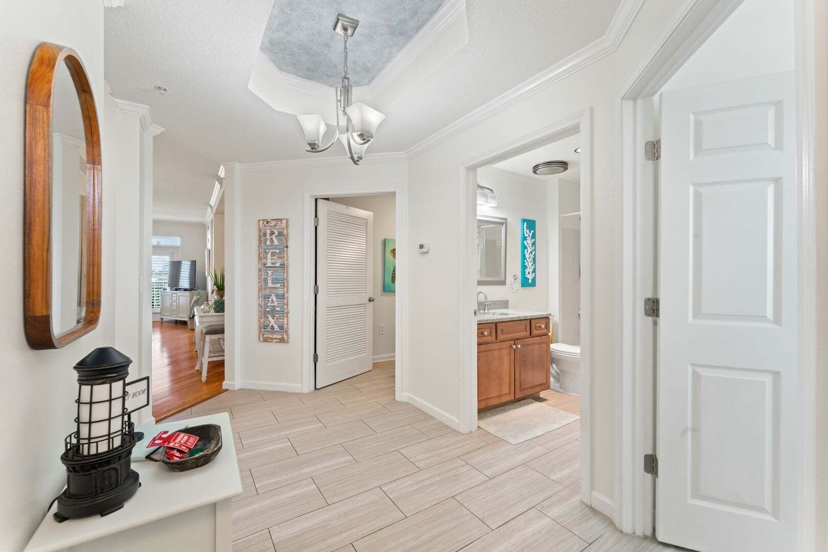 A hallway with a wooden floor, a mirror, a wall-mounted light fixture, and a door leading to a bathroom.