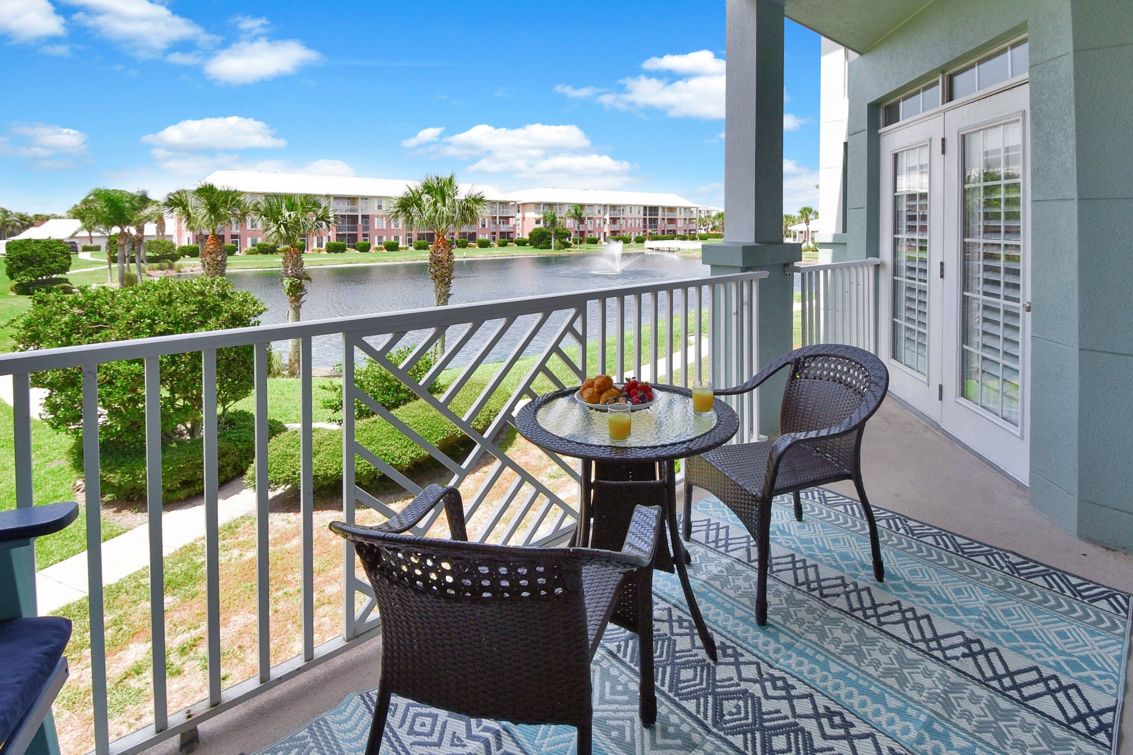 A balcony with two wicker chairs, a small round table with a fruit plate and drinks, and a view of a water body and buildings in the background.
