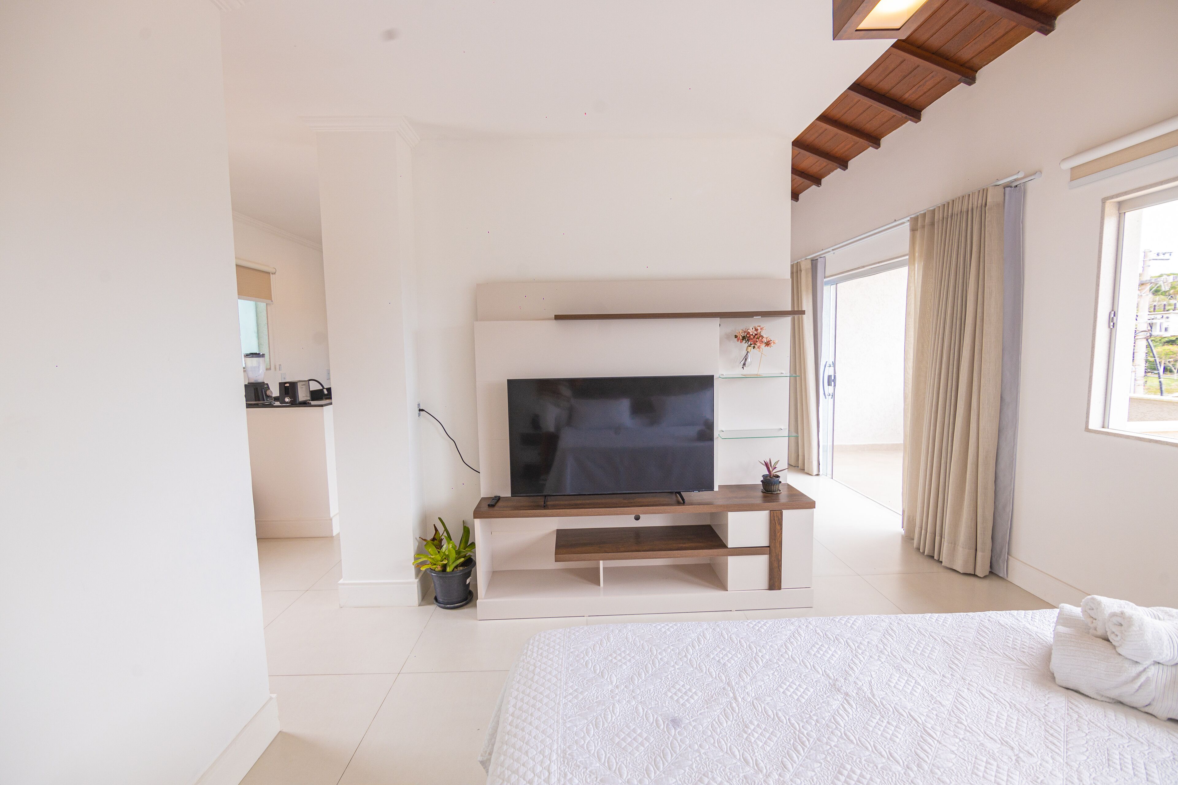 A bedroom with a bed, a TV on a wooden stand, a potted plant, and a window with curtains.