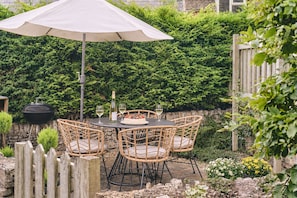 Une terrasse extérieure avec une table ronde, des chaises en osier, un barbecue et un grand parasol.