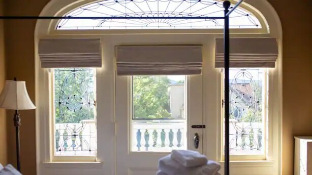 A hotel room with a bed, a lamp, a window with a view of greenery, and a door with a balcony.