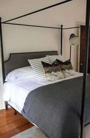 A four-poster bed with white bedding and striped pillows, a gray throw blanket, and a bedside lamp.
