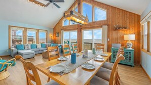 A spacious living room with a wooden dining table set for four, a sofa, and a view of the beach. - Oceanfront Dog Friendly Home w/ Hot Tub and Nearby Beach Access (Avon)