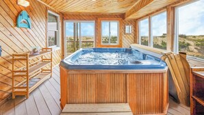 A wooden cabin interior with a hot tub, wooden deck, and large windows offering a view of the outdoors. - Oceanfront Dog Friendly Home w/ Hot Tub and Nearby Beach Access (Avon)