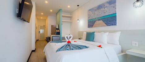 A hotel room with a bed, a painting of a beach scene, a TV mounted on the wall, and a hallway leading to a bathroom.