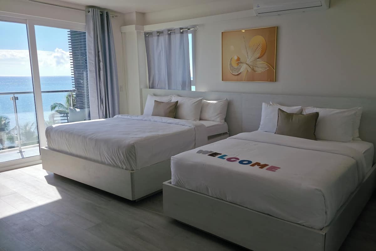 Habitación de hotel con dos camas, un ventanal con vista al océano y un cuadro en la pared.